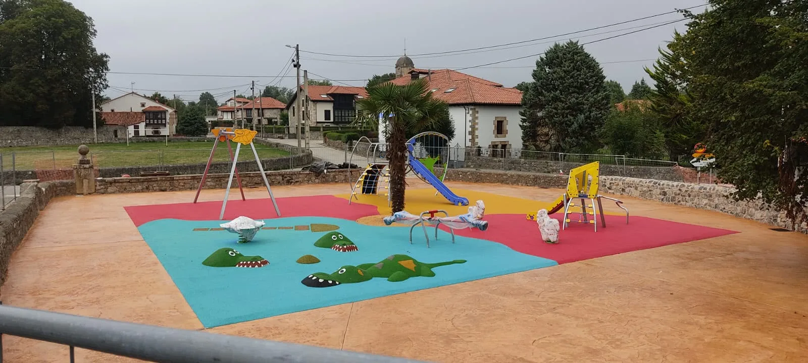 Vista general de un parque infantil con columpios, tobogán y pavimento de caucho multicolor